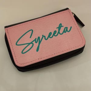 Puede incluir: Una cartera rosa con cremallera con el nombre "Syreeta" escrito en cursiva turquesa.