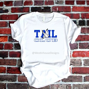 May include: White t-shirt with a blue and white graphic that says "TAIL CLUB" with a white dove flying above the word "TAIL". The text "@WestinhouseDesigns" is below the graphic.