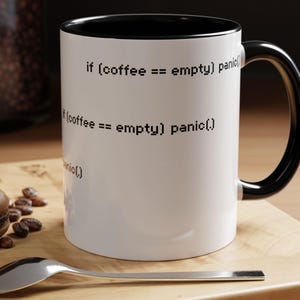May include: White coffee mug with black interior and handle. The mug features the text "if (coffee == empty) panic()" in black. A silver spoon and a macaron are on a wooden surface next to the mug.