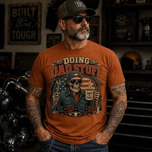May include: Orange t-shirt featuring the text "DOING DAD STUFF" and a graphic of a skeleton wearing a cap, holding a wrench and a mug that says "DAD FUEL". The shirt has a crew neck and short sleeves.