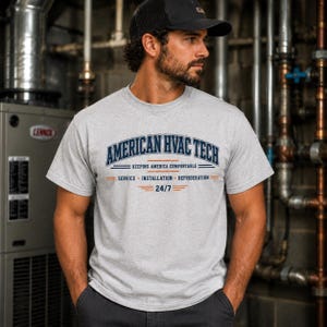 May include: A light grey short-sleeve t-shirt featuring the dark blue text AMERICAN HVAC TECH, the phrase KEEPING AMERICA COMFORTABLE, and the words SERVICE, INSTALLATION, REFRIGERATION, and 24/7 with orange horizontal accent lines.