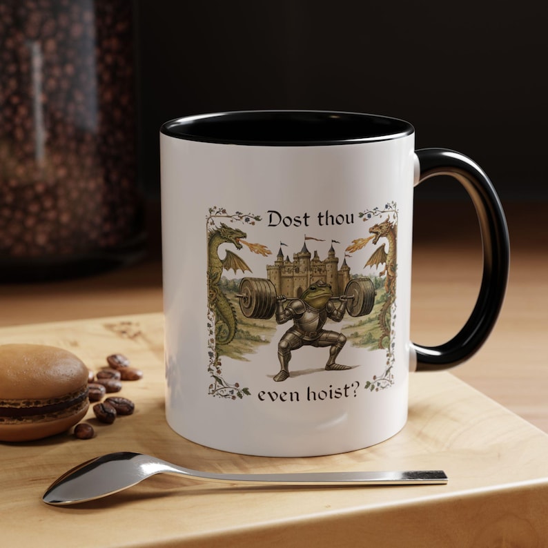 May include: White ceramic mug with a black interior and handle. The mug features a whimsical illustration of a frog in armour lifting weights, with dragons and a castle. Text reads "Dost thou even hoist?"