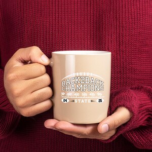 Pu&ograve; includere: Una tazza in ceramica beige con la scritta "BACK2BACK CHAMPIONS" in bianco e grigio, con una grafica di football americano. La tazza ha anche i numeri "24" e "25". La tazza &egrave; tenuta da una persona che indossa un maglione bordeaux.
