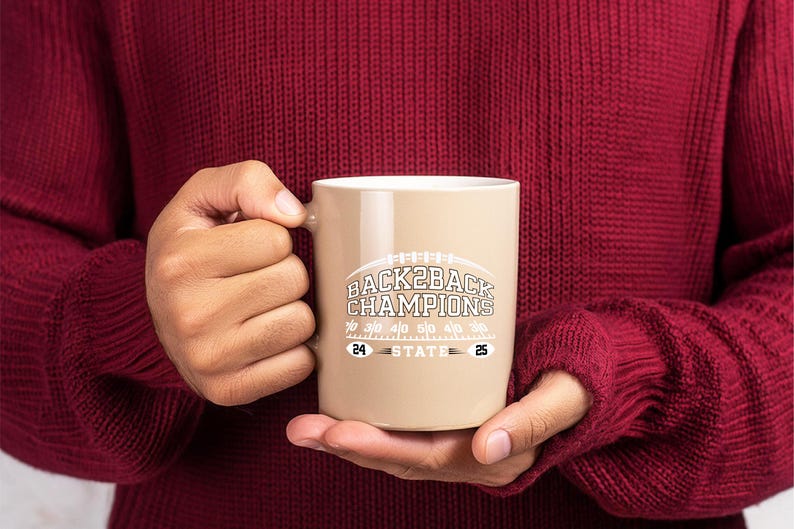 Pu&ograve; includere: Una tazza in ceramica beige con la scritta "BACK2BACK CHAMPIONS" in bianco e grigio, con una grafica di football americano. La tazza ha anche i numeri "24" e "25". La tazza &egrave; tenuta da una persona che indossa un maglione bordeaux.