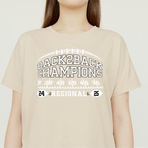 Pu&ograve; includere: T-shirt beige con la scritta bianca "BACK2BACK CHAMPIONS" sopra una grafica di campo da football. Sotto, i numeri "24" e "25" con "REGIONAL" in mezzo. La maglietta &egrave; in stile casual.