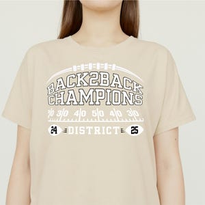 Pu&ograve; includere: Una maglietta beige con la scritta "BACK2BACK CHAMPIONS" in bianco, sopra una grafica di campo da football. La maglietta presenta anche i numeri "24 DISTRICT 25".