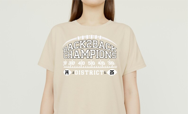 Pu&ograve; includere: Una maglietta beige con la scritta "BACK2BACK CHAMPIONS" in bianco, sopra una grafica di campo da football. La maglietta presenta anche i numeri "24 DISTRICT 25".