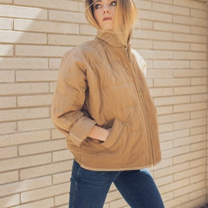 May include: A woman wearing a brown quilted jacket with a zipper closure and blue jeans. She is standing in front of a brick wall.