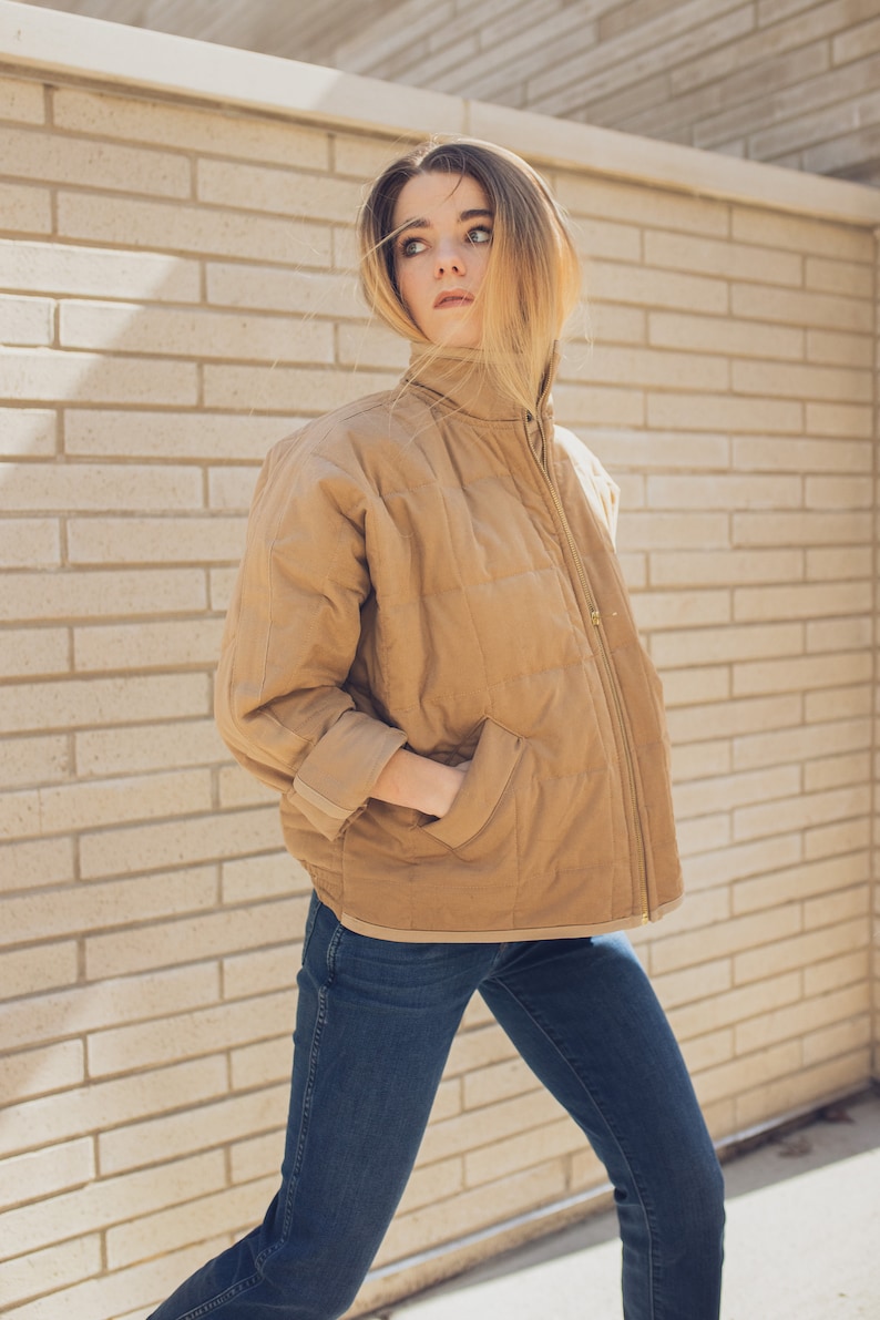 May include: A woman wearing a brown quilted jacket with a zipper closure and blue jeans. She is standing in front of a brick wall.