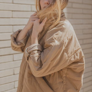 May include: A woman wearing a brown quilted jacket with a high collar. She is standing in front of a brick wall and has her hair covering her face.