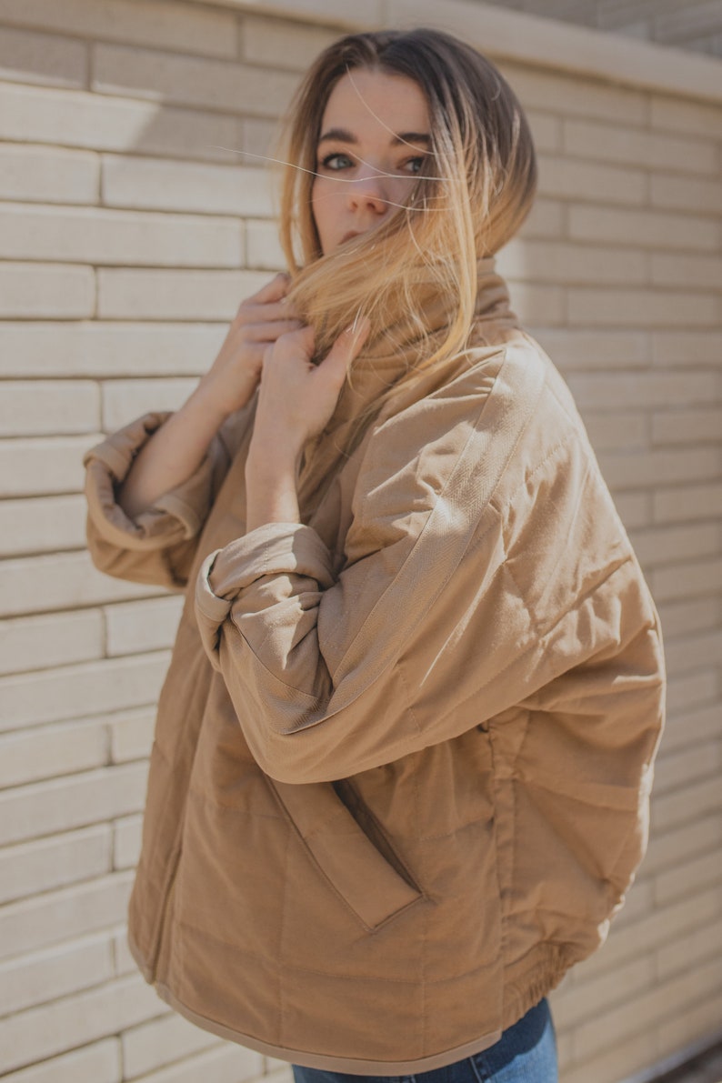 May include: A woman wearing a brown quilted jacket with a high collar. She is standing in front of a brick wall and has her hair covering her face.
