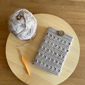 Op de afbeelding: Een beige gehaakte boekomslag met een houten knoopsluiting. Een bol beige garen en een haaknaald zijn ook op de afbeelding te zien.