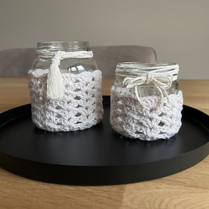 Op de afbeelding: Twee glazen potten met witte gehaakte deksels. De potten staan op een zwarte ronde dienblad. De grotere pot heeft een witte kwast die van boven hangt.