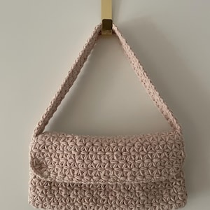 Op de afbeelding: Een lichtroze, handgemaakte gehaakte handtas met een geometrisch patroon. De tas heeft een rechthoekige body met een klepsluiting en een gevlochten band. De tas hangt aan een goudkleurige haak.