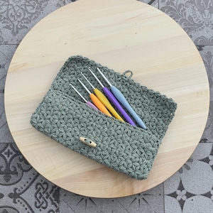 Op de afbeelding: Een saliegroene haaknaald etui met een houten knevelsluiting, met daarin verschillende kleurrijke haaknaalden. De etui ligt op een licht houten rond oppervlak, tegen een grijze achtergrond met patronen.