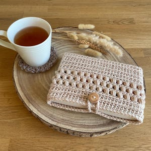 Op de afbeelding: Een beige gehaakte boekomslag met een houten knoopsluiting ligt op een houten schijf, naast een witte theemok en een bijpassende onderzetter. Gedroogd siergras voegt een natuurlijk tintje toe.