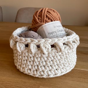 Op de afbeelding: Een witte gehaakte mand met een gegolfde rand, gevuld met twee bollen garen, een bruine en een oranje. Het garen is gelabeld "Happy Chunky" en is een zacht, dik garen, perfect om te haken.