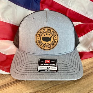May include: A grey and black trucker hat featuring a circular wooden patch on the front. The patch displays a black silhouette of the United States map, three stars above and below, and the text LAND OF THE FREE HOME OF THE BRAVE. A black sticker on the brim reads EST. 1970 RICHARDSON ORIGINAL 112 TRUCKER ONE SIZE.
