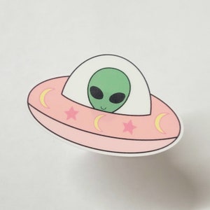 May include: A pink and white cartoon UFO with a green alien inside. The UFO has yellow crescent moons and pink stars on the side.