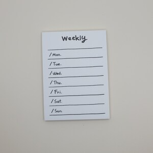 May include: A white weekly planner notepad with black lines and the word "Weekly" written at the top. The days of the week are listed on the left side of the notepad: Monday, Tuesday, Wednesday, Thursday, Friday, Saturday, and Sunday.