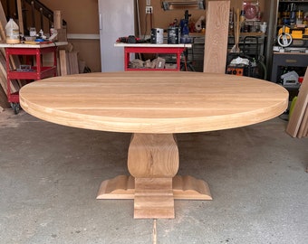 Könnte beinhalten: Ein großer runder Esstisch aus hellem Holz mit sichtbarer Maserung. Der Tisch verfügt über eine dicke, kreisförmige Tischplatte, die von einem zentralen Säulenfuß getragen wird, der sich nach unten hin zu einem breiten, gestuften Sockel verbreitert.