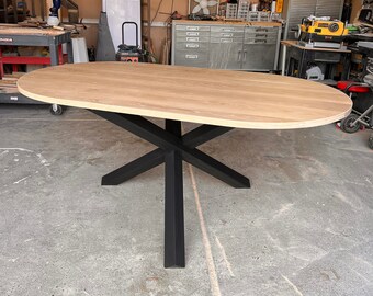 Könnte beinhalten: Ein ovaler Esstisch mit einer hellen, natürlich gemaserten Holzplatte und glatter Oberfläche. Der Tisch wird von einem zentralen Untergestell getragen, das aus vier sich kreuzenden schwarzen Metallbeinen in X-Form besteht.