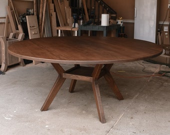 Könnte beinhalten: Ein großer, runder Esstisch aus dunkelbraunem Holz mit einer glatten, kreisförmigen Tischplatte. Der Tisch verfügt über ein zentrales Gestell mit vier schrägen Beinen, die in der Mitte zusammenlaufen und durch eine kleine quadratische Ablage verbunden sind.