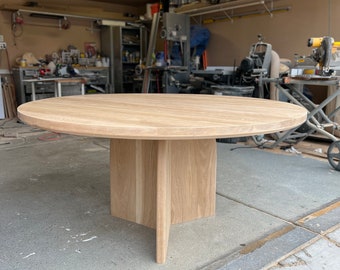 Könnte beinhalten: Ein runder Tisch aus hellem Holz mit einer glatten, kreisförmigen Tischplatte und einem zentralen Sockel, der aus drei sich kreuzenden vertikalen Paneelen besteht, die in einer Werkstatt für Stabilität sorgen.