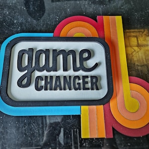 May include: A vibrant wooden sign featuring the text "game changer" in black, decorative font. The sign has a rectangular shape with a blue border and a white interior. It also includes a retro-style rainbow and striped design.