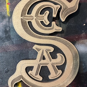 May include: A wooden cutout featuring the letters S, C, and A intertwined in a decorative, serif-style font. The surface has a light tan, natural wood grain finish with dark, engraved outlines and recessed details that create a layered appearance.