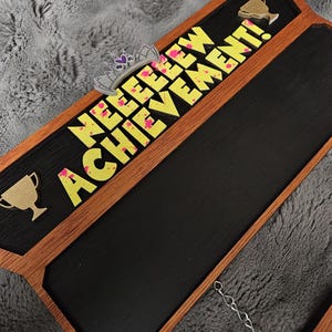May include: A rectangular wooden sign with a black centre panel featuring the yellow text "NEEEEEW ACHIEVEMENT!" with pink paint splatters. The sign includes a silver tiara cutout, two gold trophy cutouts, and a small silver metal hanging chain.