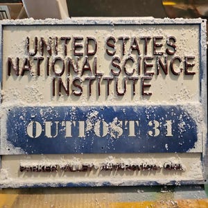 May include: A rectangular sign with the text "UNITED STATES NATIONAL SCIENCE INSTITUTE" in raised, dark red letters. Below, a blue section reads "OUTPOST 31" in white. The sign is covered in a white, textured material.