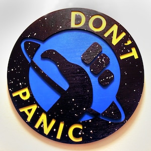 Può includere: Un cartello rotondo in legno nero e blu con le parole "DON'T PANIC" in lettere gialle. Il cartello presenta un'astronave stilizzata con un gesto di pollice in su.