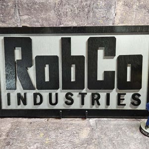 May include: Black framed sign with the text "RobCo INDUSTRIES" in black letters on a silver background. A small Vault-Tec figurine stands to the right of the sign. The sign has two small hooks at the bottom.