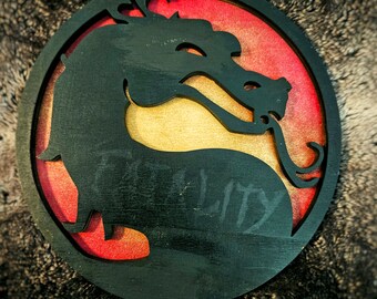 Könnte beinhalten: Ein rundes Schild aus schwarzem Holz mit rotem und gelbem Hintergrund. Das Schild zeigt eine schwarze Silhouette eines Drachen mit dem Wort "Mortal" in Schwarz.