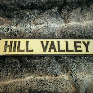 Puede incluir: Un letrero de madera con letras negras que dice "HILL VALLEY". El letrero está sobre una superficie peluda marrón.