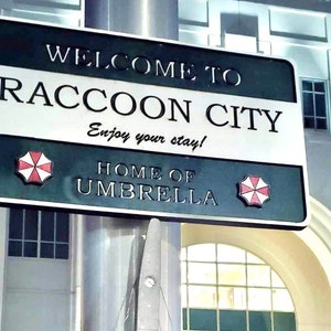 May include: A black and green sign with white lettering that reads "Welcome to Raccoon City, Enjoy your stay! Home of Umbrella". The sign has two red and white umbrella logos on either side.