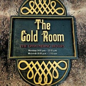 May include: A black wooden sign with gold trim and lettering. The sign reads "The Gold Room" and lists the "Unwinding Hours" as Weekdays 9:00 pm - 12:00 pm and Weekends 8:00 pm - 1:00 am.