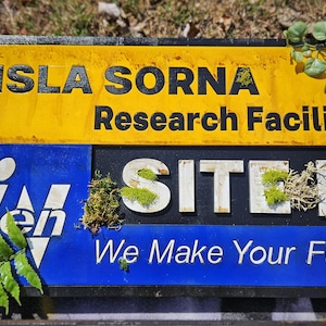 Może przedstawiać: Żółto-niebieski znak z napisem "Isla Sorna Research Facility Site B We Make Your Future" z zielonym mchem rosnącym na znaku.