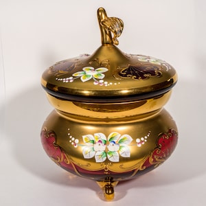 May include: A decorative, gold-coloured jar with a lid, featuring hand-painted floral designs in white, yellow, and red. The jar has a bulbous base and a lid with a decorative handle. The jar is approximately 20 cm tall.
