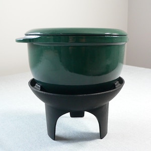 Op de afbeelding: Een donkergroene geëmailleerde gietijzeren braadpan met bijpassend deksel, rustend op een zwarte gietijzeren driepoot. De pot heeft een afgeronde vorm en zijhandgrepen. Een klassieker om in te koken.