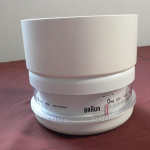 Puede incluir: Balanza de cocina Braun blanca con un cuenco medidor circular transparente. La báscula tiene un dial con medidas en onzas y kilogramos, con una capacidad máxima de 2,04 kg. Diseñada para medir ingredientes.