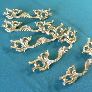 May include: Five ornate brass drawer pulls with a decorative floral design. The pulls are arranged in a row on a blue surface.