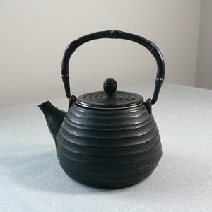 May include: A black cast iron teapot with a bamboo-style handle and a matching lid. The teapot has a rounded body with horizontal ridges and a spout for pouring. The handle is arched, and the lid has a small knob.
