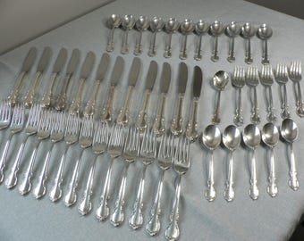 Può includere: Una collezione di posate color argento, tra cui forchette, coltelli e cucchiai, disposte su una superficie grigio chiaro. Le posate presentano dettagli decorati sul manico. Il set include diverse posate, ideale per un'occasione formale.