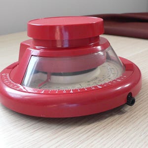 May include: A red vintage timer with a clear plastic dome and a circular dial. The timer has a round base and a top dial. The dial has white numbers and red markings. The timer is on a light wood surface.