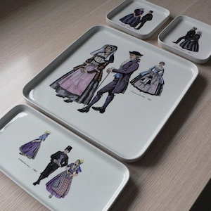 May include: A collection of four white ceramic trays, each adorned with illustrations of figures in historical attire. The trays are in various sizes and shapes, including rectangular and square. The illustrations depict people in colourful clothing, evoking a historical theme.
