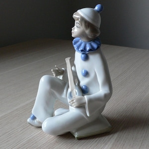 Könnte beinhalten: Eine Porzellanfigur eines Clowns, der ein Musikinstrument spielt. Der Clown trägt ein weißes und blaues Kostüm mit einem blauen Hut. Die Figur sitzt auf einem Sockel.
