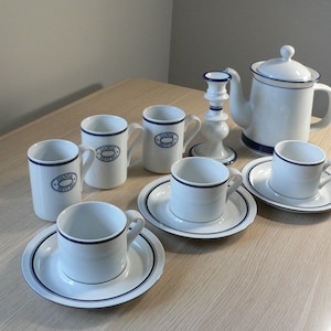 May include: A white ceramic tea set with blue detailing, including a teapot, cups, saucers, and mugs. The mugs feature the text "Bistro". A white candlestick holder is also present.