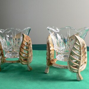 May include: Two clear glass candle holders with a decorative leaf design on the metal base. The holders are on a green surface.
